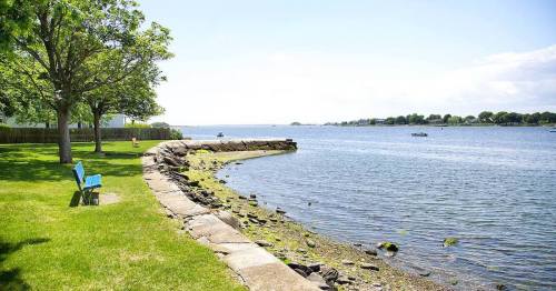 A serene waterfront scene with a grassy area, a stone wall, and blue chairs overlooking the calm water and distant boats.