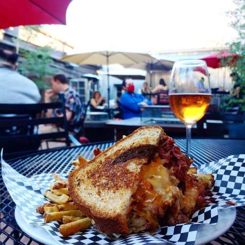 A close-up of a grilled sandwich with melted cheese and fries, with a glass of rosé in a lively outdoor setting.