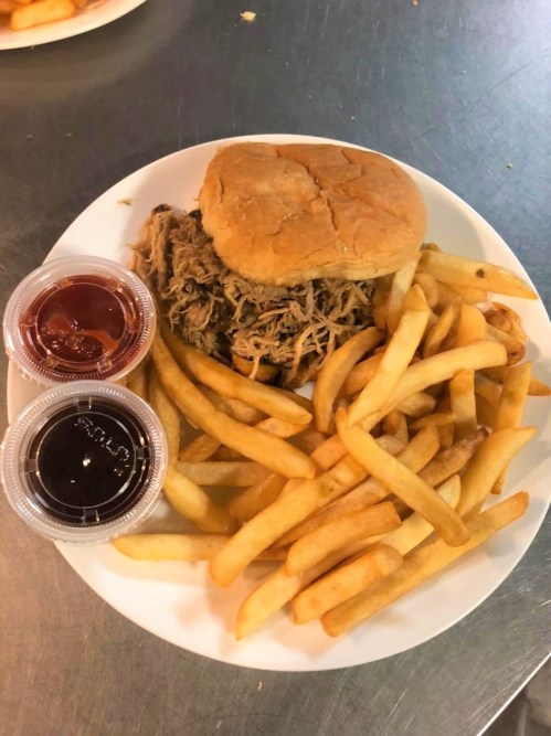 A plate with a pulled pork sandwich, golden fries, and two dipping sauces.