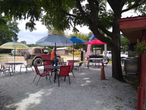 Outdoor seating area with colorful umbrellas, tables, and a parked vehicle under trees, featuring an American flag.