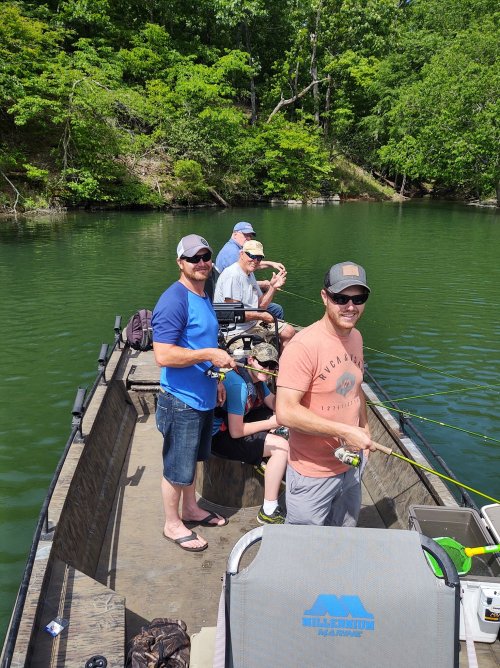 A group of five people fishing from a boat on a calm lake surrounded by greenery.