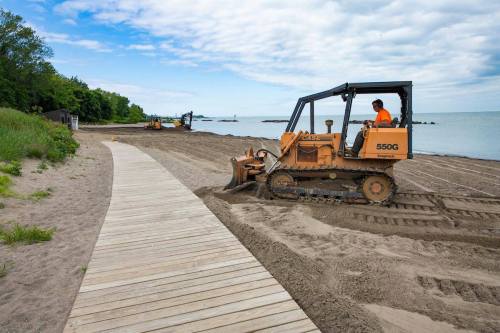 A bulldozer levels sand on a beach, with a wooden walkway and lake in the background under a cloudy sky.
