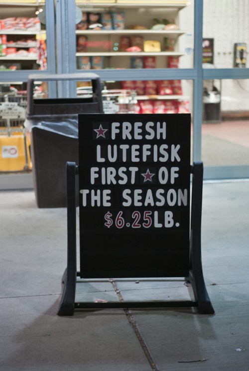 Sign outside a store advertising fresh lutefisk for $6.25 per pound, with a display of products in the background.