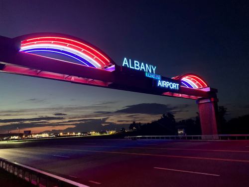 Neon-lit sign for Albany International Airport at dusk, with colorful arches and a clear sky in the background.