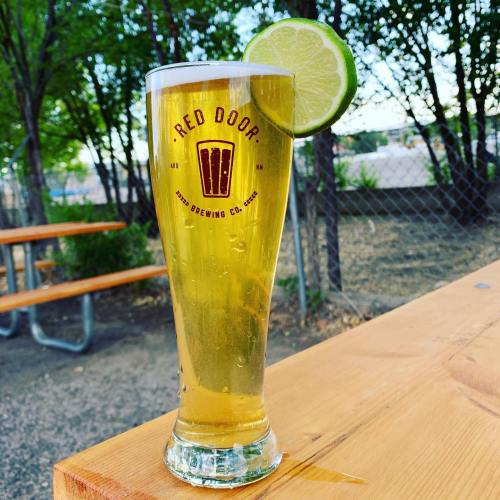 A tall glass of beer with a lime wedge, featuring the logo of Red Door Brewing Co., on a wooden table outdoors.
