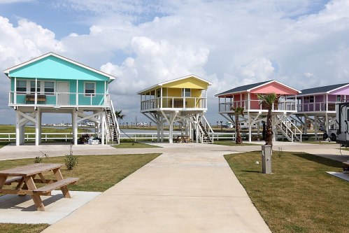Colorful beach houses on stilts line a pathway, surrounded by green grass and palm trees under a cloudy sky.