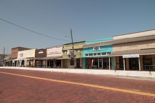 A row of colorful storefronts along a brick-paved street under a clear blue sky.
