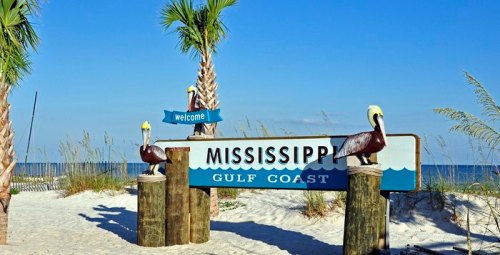 Welcome sign for Mississippi Gulf Coast, featuring pelican sculptures and palm trees against a clear blue sky.