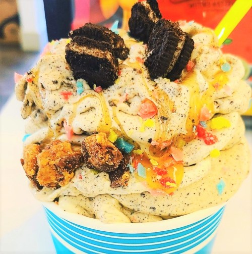 A colorful ice cream cup topped with crushed cookies, caramel drizzle, and sprinkles.