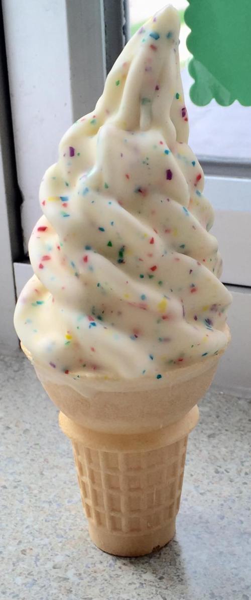 A soft-serve ice cream cone with colorful sprinkles on a light-colored swirl, set against a window.