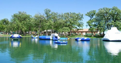 Inflatable water toys on a calm lake surrounded by green trees and a clear blue sky.