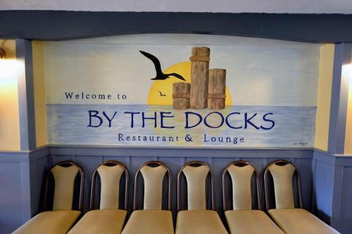 Mural of a sunset over water with the text "Welcome to By The Docks Restaurant & Lounge" and empty chairs below.