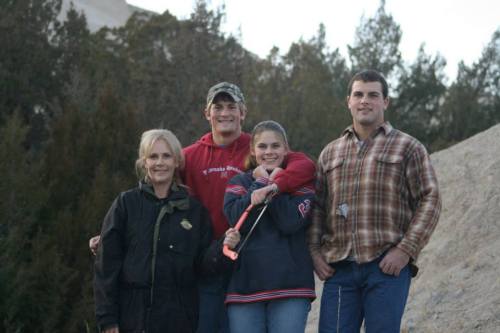 A group of four people stands together outdoors, smiling, with trees and a hillside in the background.