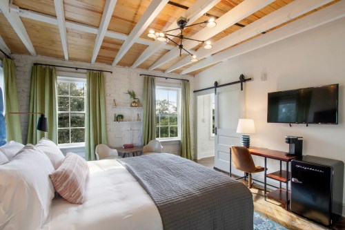 Cozy bedroom with a bed, TV, desk, and mini fridge, featuring wooden beams and large windows with green curtains.