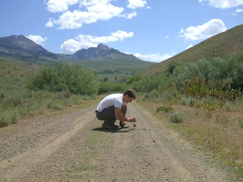 A person crouches on a dirt path, photographing the ground with mountains and greenery in the background.