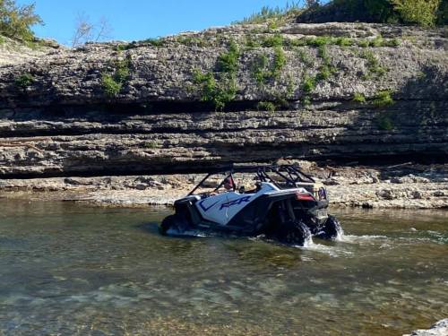 A side-by-side vehicle driving through shallow water near rocky terrain under a clear blue sky.