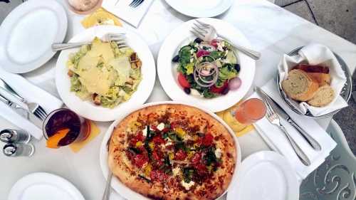 A table set with a salad, pizza topped with vegetables, and bread, accompanied by drinks in glasses.