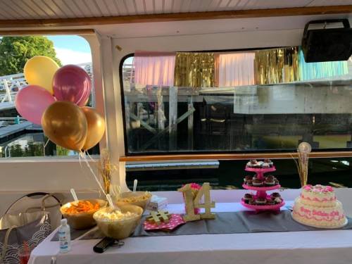 A festive table on a boat with balloons, snacks, and a pink cake, decorated for a celebration.
