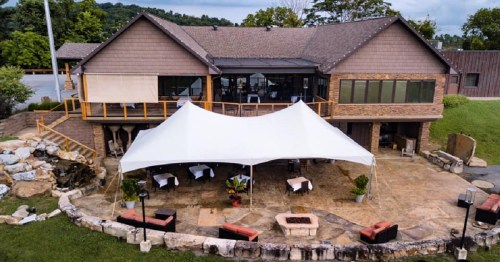 A spacious outdoor venue with a large white tent, stone patio, and a modern building surrounded by greenery.