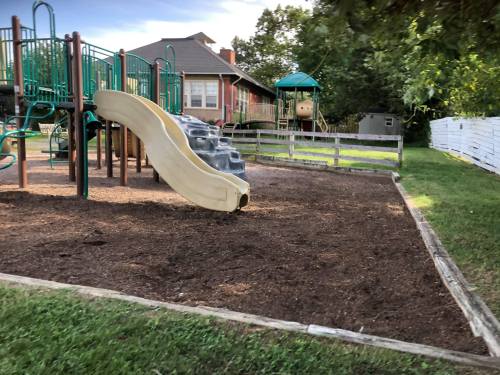 A playground with a slide, climbing structures, and a gazebo in a grassy area surrounded by trees and a fence.
