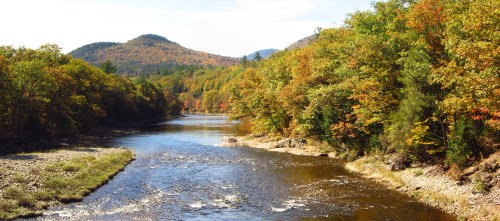 A serene river flows through a landscape of vibrant autumn trees and distant mountains under a clear blue sky.