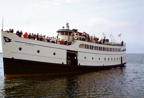 A vintage boat named Quonset with passengers on deck, sailing on calm waters under a clear sky.