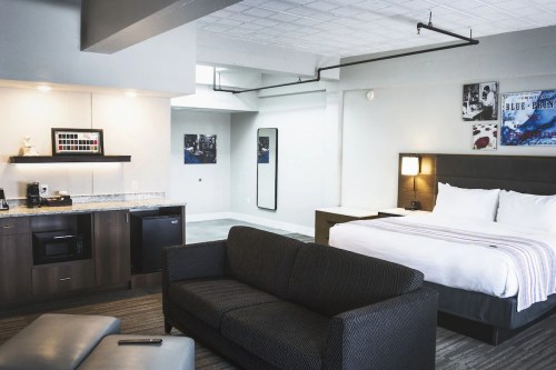 A modern hotel room featuring a bed, sofa, kitchenette, and artwork on the walls. Bright and spacious design.