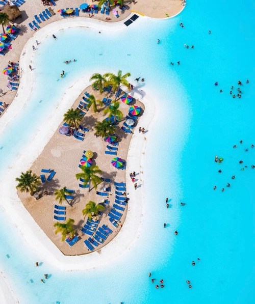 Aerial view of a vibrant pool area with umbrellas, lounge chairs, and people swimming in clear blue water.