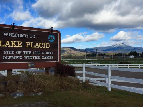 Sign welcoming visitors to Lake Placid, with mountains and a cloudy sky in the background. Site of the Olympic Winter Games.