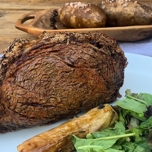 A beautifully roasted piece of meat with herbs, served alongside baked potatoes and fresh greens on a white plate.