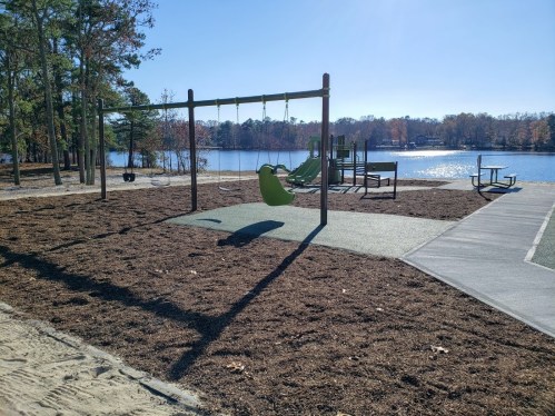 A playground with swings and a slide near a lake, surrounded by trees and a clear blue sky.