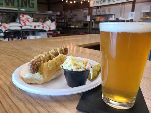 A plate with a sausage in a bun, coleslaw, pickles, and a glass of beer on a wooden table in a brewery.
