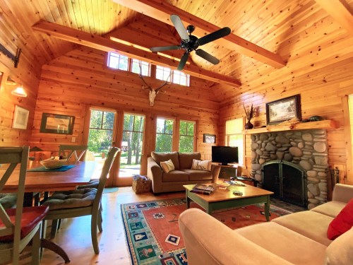 Cozy wooden cabin interior with a stone fireplace, large windows, and rustic furniture. Bright and inviting atmosphere.