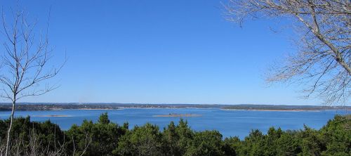 A serene lake view surrounded by trees under a clear blue sky, with distant hills on the horizon.