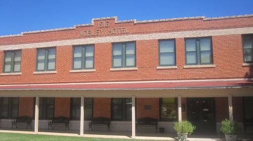 Historic red brick building with "Wobley Hotel" sign, featuring multiple windows and a covered porch. Clear blue sky above.