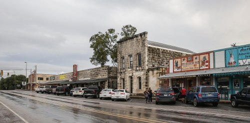 Historic buildings line a wet street, with parked cars and colorful storefronts under a cloudy sky.