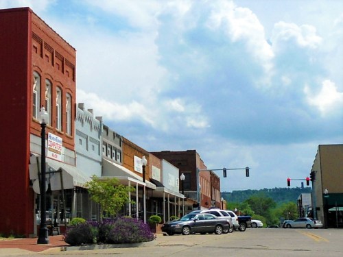 A small town street lined with brick buildings, shops, and parked cars under a cloudy sky.