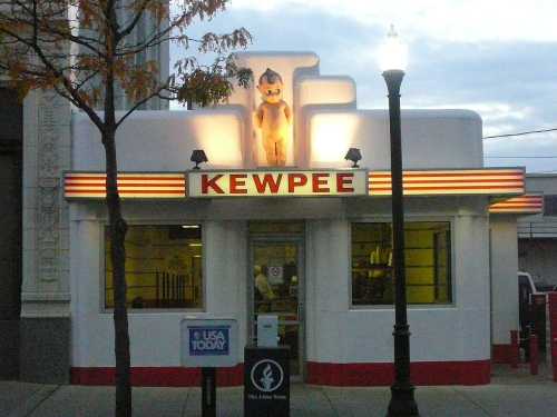 A retro diner named "Kewpee" with a cartoon figure on top, featuring bright signage and a classic architectural style.