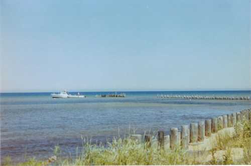 A calm shoreline with a boat near a wooden pier, surrounded by blue water and clear skies.