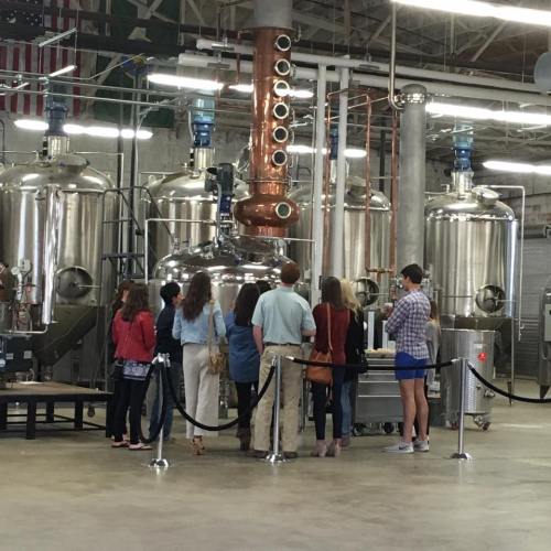 A group of people observing a distillation setup in a brewery, with large metal tanks and copper equipment in the background.