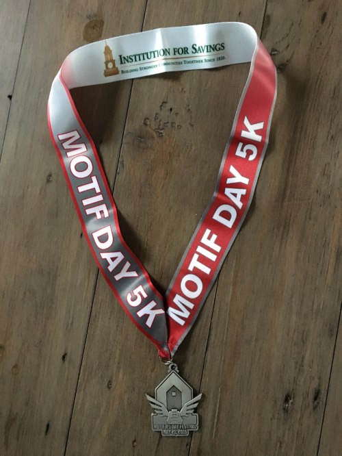 A silver medal with a ribbon reading "MOTIF DAY 5K" and "INSTITUTION FOR SAVINGS" on a wooden surface.