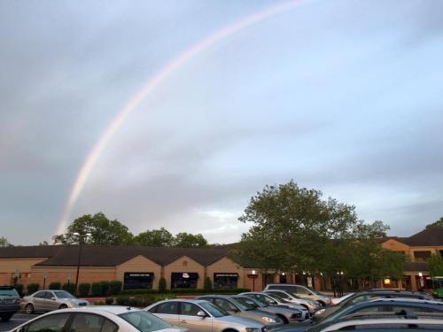 A rainbow arcs over a parking lot filled with cars, with trees and buildings in the background under a cloudy sky.