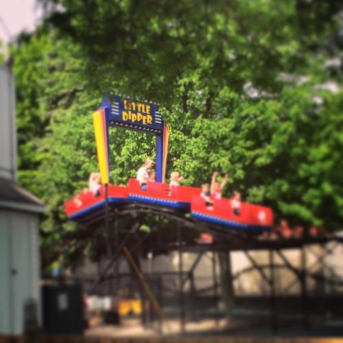 A colorful roller coaster named "Little Dipper" with riders enjoying the ride amidst green trees.