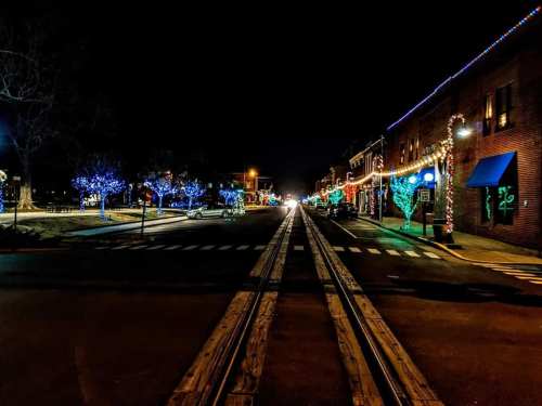 A nighttime street scene adorned with colorful holiday lights on trees and buildings, with a view of railroad tracks.