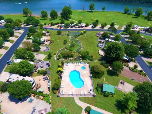 Aerial view of a campground with a pool, green lawns, trees, and a river in the background.