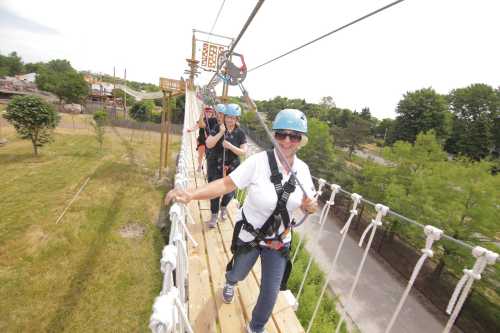 A group of people wearing helmets and harnesses walk across a suspension bridge in a scenic outdoor setting.