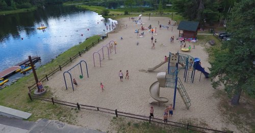 A sandy beach area with a playground, people swimming, and kayaks on a lake surrounded by trees.