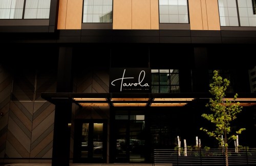 Modern building facade featuring a large sign that reads "Tavola" with greenery in front.