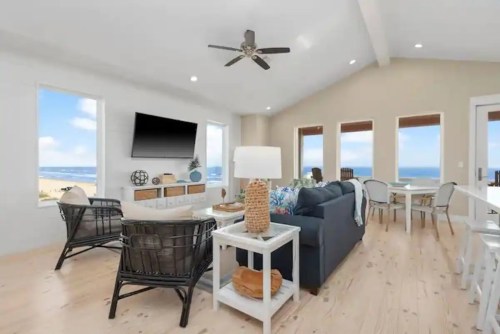 Bright coastal living room with large windows, ocean views, modern furniture, and a ceiling fan.