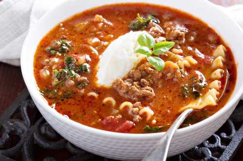 A bowl of hearty soup with ground meat, pasta, greens, and a dollop of cream, garnished with a basil leaf.